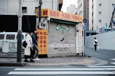 An urban street scene featuring a closed shop with a corrugated metal shutter displaying 'BEST SERVICE' in faded lettering. The shop is surrounded by buildings, and there are red, yellow, and white signs with Japanese text. Two individuals are walking on the sidewalk next to the shop, and another person is walking away on the crosswalk. The street is quiet with minimal traffic.