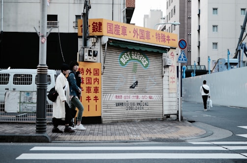 An urban street scene featuring a closed shop with a corrugated metal shutter displaying 'BEST SERVICE' in faded lettering. The shop is surrounded by buildings, and there are red, yellow, and white signs with Japanese text. Two individuals are walking on the sidewalk next to the shop, and another person is walking away on the crosswalk. The street is quiet with minimal traffic.