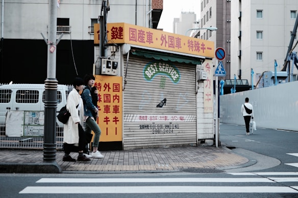 An urban street scene featuring a closed shop with a corrugated metal shutter displaying 'BEST SERVICE' in faded lettering. The shop is surrounded by buildings, and there are red, yellow, and white signs with Japanese text. Two individuals are walking on the sidewalk next to the shop, and another person is walking away on the crosswalk. The street is quiet with minimal traffic.