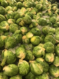 A large pile of fresh brussels sprouts, piled on a bed of crushed ice. The vegetables are bright green and glistening, indicating freshness and moisture.