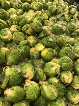 A large pile of fresh brussels sprouts, piled on a bed of crushed ice. The vegetables are bright green and glistening, indicating freshness and moisture.