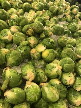 A large pile of fresh brussels sprouts, piled on a bed of crushed ice. The vegetables are bright green and glistening, indicating freshness and moisture.