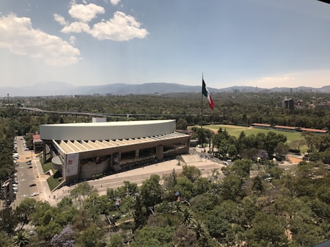 A large modern building with a distinctive architectural design is surrounded by lush greenery. In the foreground, trees fill the landscape, while a prominent flagpole with a large flag stands next to the building. The background features a sprawling urban landscape with distant mountains under a partly cloudy sky.