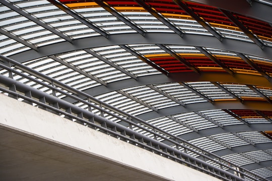 Curved metal beams and supporting structures form a canopy with alternating clear and tinted panels. The structure features red and amber hues, adding a vibrant contrast to the metallic tones.