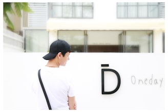 A person wearing a black cap is facing away from the camera, standing in front of a white wall with a large letter 'D' and the word 'Oneday'. The background includes a building with large windows and some greenery.