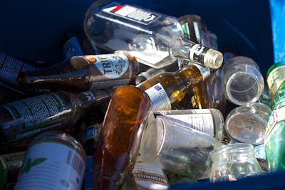 A collection of empty glass bottles is scattered inside a blue recycling bin. Various bottle shapes and sizes are visible, with labels on some bottles including text and graphics. The sunlight casts reflections and highlights on the glass surfaces, adding a mix of shadows.