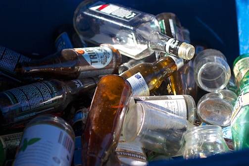 A collection of empty glass bottles is scattered inside a blue recycling bin. Various bottle shapes and sizes are visible, with labels on some bottles including text and graphics. The sunlight casts reflections and highlights on the glass surfaces, adding a mix of shadows.