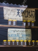Two signs advertising natural honey are displayed. The top sign features Japanese text with smiley faces and weathered texture, while the bottom sign has text in English and Urdu, depicting mountains. Below each sign are rows of small glass jars with yellow lids.