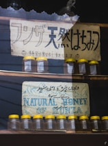 Two signs advertising natural honey are displayed. The top sign features Japanese text with smiley faces and weathered texture, while the bottom sign has text in English and Urdu, depicting mountains. Below each sign are rows of small glass jars with yellow lids.