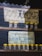 Two signs advertising natural honey are displayed. The top sign features Japanese text with smiley faces and weathered texture, while the bottom sign has text in English and Urdu, depicting mountains. Below each sign are rows of small glass jars with yellow lids.