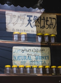 Two signs advertising natural honey are displayed. The top sign features Japanese text with smiley faces and weathered texture, while the bottom sign has text in English and Urdu, depicting mountains. Below each sign are rows of small glass jars with yellow lids.