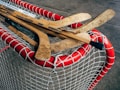 Several wooden hockey sticks are placed on top of a red and white hockey goal net. The sticks appear worn and used, with visible scratches and markings. The netting is tightly woven, and the background shows a concrete surface.