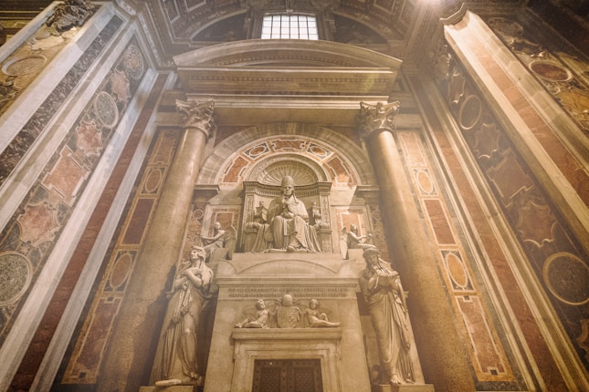 A grand interior space featuring a detailed stone sculpture of a seated pope or religious figure adorned with intricate carvings. The scene is framed by ornate columns and elaborate frescoes with historical designs decorating the surrounding walls.