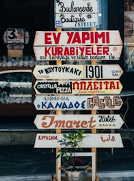 A collection of various colorful signs with different fonts and languages is displayed in front of a shop. The signs advertise different types of businesses, such as bakeries, hotels, and pizzerias. Below the signs is a small plant in a metal bucket with a label that reads 'Flowers.'