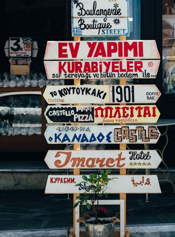 A collection of various colorful signs with different fonts and languages is displayed in front of a shop. The signs advertise different types of businesses, such as bakeries, hotels, and pizzerias. Below the signs is a small plant in a metal bucket with a label that reads 'Flowers.'