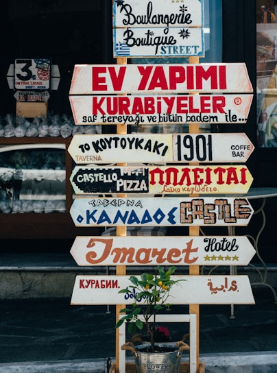 A collection of various colorful signs with different fonts and languages is displayed in front of a shop. The signs advertise different types of businesses, such as bakeries, hotels, and pizzerias. Below the signs is a small plant in a metal bucket with a label that reads 'Flowers.'