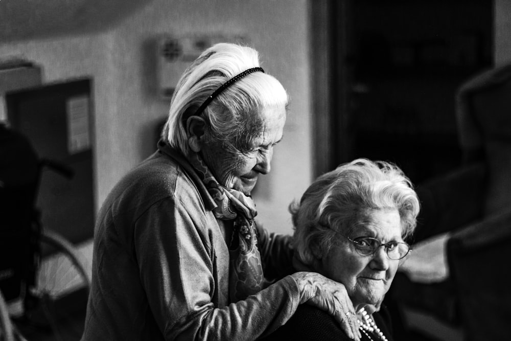 A standing elderly woman rests her hands on the shoulders of another woman
