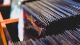 Close-up view of a stack of vinyl records lined up vertically, some partially visible through clear protective sleeves. The scene is softly lit, creating a nostalgic and cozy atmosphere.