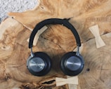 A pair of sleek, black over-ear headphones rests on a textured wooden surface with visible grains and knots. The headphones have round ear cups and a padded headband, displaying a metallic logo on the outer side of the ear cups.