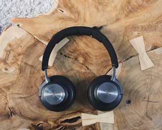 A pair of sleek, black over-ear headphones rests on a textured wooden surface with visible grains and knots. The headphones have round ear cups and a padded headband, displaying a metallic logo on the outer side of the ear cups.