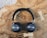 A pair of sleek, black over-ear headphones rests on a textured wooden surface with visible grains and knots. The headphones have round ear cups and a padded headband, displaying a metallic logo on the outer side of the ear cups.