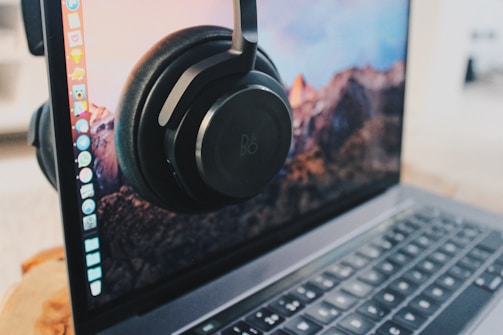 A pair of headphones is placed on the screen of an open laptop. The screen displays a scenic mountain landscape as the desktop wallpaper, with several application icons on the left side. The laptop is on a wooden surface.