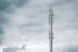 A tall communication tower reaches into a cloudy sky, with several antennas mounted on its structure. The clouds appear thick and grey, suggesting an overcast day.