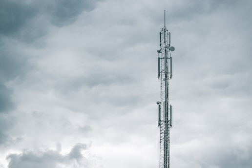 A tall communication tower reaches into a cloudy sky, with several antennas mounted on its structure. The clouds appear thick and grey, suggesting an overcast day.