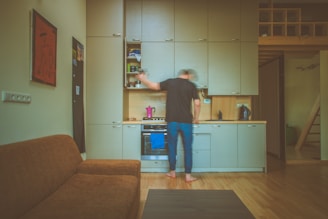 A person with blurred motion is standing in a compact kitchen area, reaching into one of the overhead cabinets. The kitchen is equipped with light-colored cabinetry, a stove, and a kettle. A brown couch is visible in the foreground, adjacent to a wall with some framed artwork and a bulletin board. The interior is warmly lit and has wooden floors.