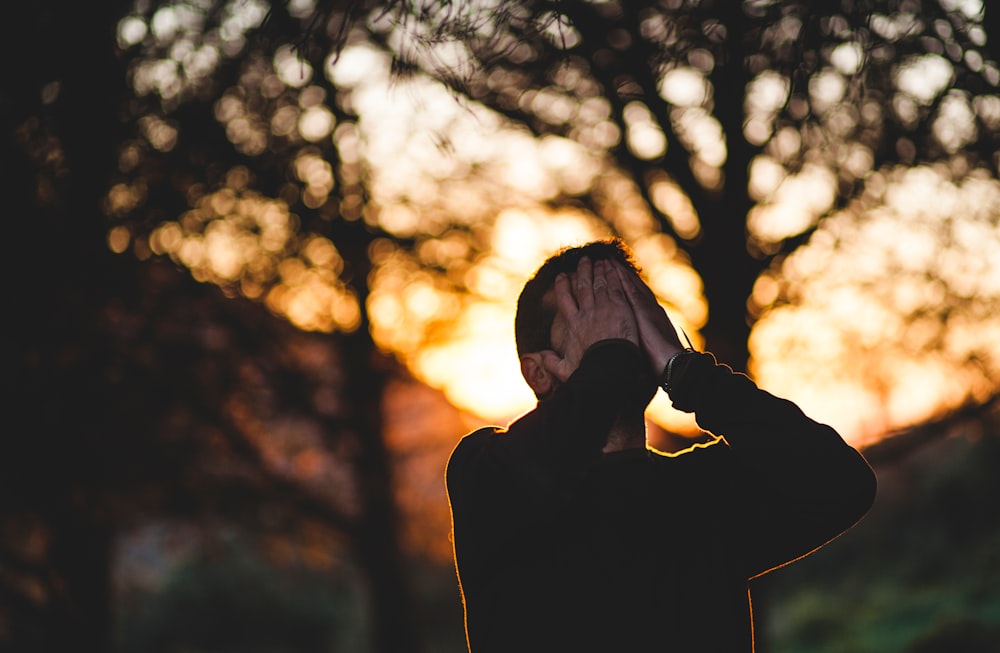 A man holds his hands over his face in front of trees at sunset