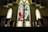 A church interior featuring large stained glass windows with vibrant colors. The central window depicts a religious figure, possibly Jesus, surrounded by nature with trees and a river path. Wooden pews line the foreground, creating a symmetrical layout leading towards the windows.