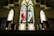 A church interior featuring large stained glass windows with vibrant colors. The central window depicts a religious figure, possibly Jesus, surrounded by nature with trees and a river path. Wooden pews line the foreground, creating a symmetrical layout leading towards the windows.