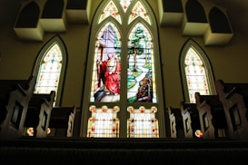 A church interior featuring large stained glass windows with vibrant colors. The central window depicts a religious figure, possibly Jesus, surrounded by nature with trees and a river path. Wooden pews line the foreground, creating a symmetrical layout leading towards the windows.