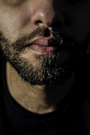 A close-up of the lower half of a person's face, showing a well-groomed, thick beard and moustache. The lips are slightly closed, and the skin appears textured. The lighting is dim, creating shadows and emphasizing the contours of the person's face.