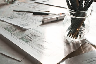 A cluttered workspace featuring several sheets of paper with sketches and diagrams spread out on a table. A jar contains multiple pencils and pens, and a marker and ruler are also present. Natural light filters through, casting shadows over the objects.
