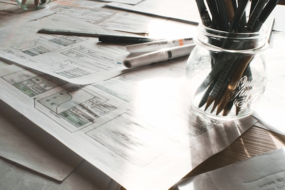 A cluttered workspace featuring several sheets of paper with sketches and diagrams spread out on a table. A jar contains multiple pencils and pens, and a marker and ruler are also present. Natural light filters through, casting shadows over the objects.