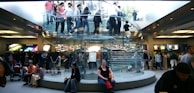A bustling retail store with a modern, transparent spiral staircase at its center. Numerous people are seen on the staircase and throughout the store, some carrying shopping bags. Bright product displays line the walls, showing colorful graphics. The environment is lively and busy.
