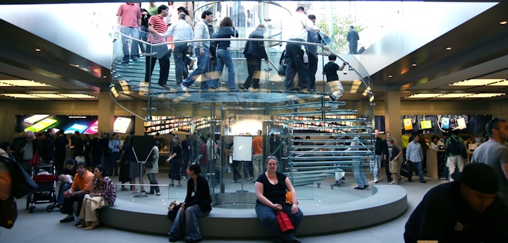A bustling retail store with a modern, transparent spiral staircase at its center. Numerous people are seen on the staircase and throughout the store, some carrying shopping bags. Bright product displays line the walls, showing colorful graphics. The environment is lively and busy.