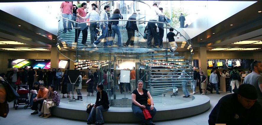 A bustling retail store with a modern, transparent spiral staircase at its center. Numerous people are seen on the staircase and throughout the store, some carrying shopping bags. Bright product displays line the walls, showing colorful graphics. The environment is lively and busy.