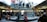 A bustling retail store with a modern, transparent spiral staircase at its center. Numerous people are seen on the staircase and throughout the store, some carrying shopping bags. Bright product displays line the walls, showing colorful graphics. The environment is lively and busy.