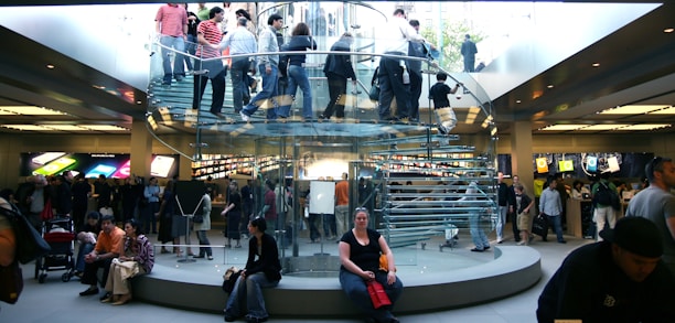 A bustling retail store with a modern, transparent spiral staircase at its center. Numerous people are seen on the staircase and throughout the store, some carrying shopping bags. Bright product displays line the walls, showing colorful graphics. The environment is lively and busy.
