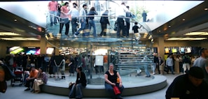 A bustling retail store with a modern, transparent spiral staircase at its center. Numerous people are seen on the staircase and throughout the store, some carrying shopping bags. Bright product displays line the walls, showing colorful graphics. The environment is lively and busy.