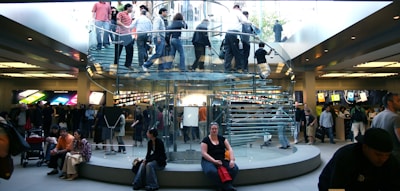 A bustling retail store with a modern, transparent spiral staircase at its center. Numerous people are seen on the staircase and throughout the store, some carrying shopping bags. Bright product displays line the walls, showing colorful graphics. The environment is lively and busy.