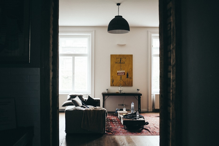 A cozy, dimly lit room with large windows allowing soft natural light to enter. The room features a comfortable sofa with cushions and a throw blanket, placed on a wooden floor. A large piece of artwork hangs on the wall, and there is a table under it with some objects. A red patterned carpet adds warmth to the space. The overall atmosphere is calm and inviting.