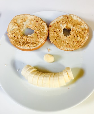 A playful arrangement of food resembling a smiling face is on a white plate. Two peanut-butter-covered bagel halves form the eyes, with a slice of banana as the nose and a series of banana slices making a curved line for the mouth.