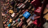 A dense collection of colorful padlocks attached to a metal fence, symbolizing committed relationships. The locks vary in color, with some having engraved messages or decorations.