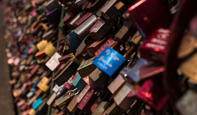 A dense collection of colorful padlocks attached to a metal fence, symbolizing committed relationships. The locks vary in color, with some having engraved messages or decorations.