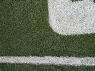 Close-up of a section of a football field showing green artificial turf with white painted lines. The texture of the grass and the distinct boundary markings are visible.