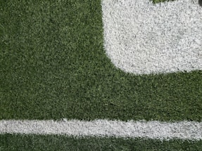 Close-up of a section of a football field showing green artificial turf with white painted lines. The texture of the grass and the distinct boundary markings are visible.