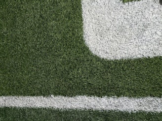 Close-up of a section of a football field showing green artificial turf with white painted lines. The texture of the grass and the distinct boundary markings are visible.